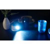 Kleines rundes Leuchtgerät strahlt starkes blaues Licht über eine geriffelte Oberfläche; rechts ein durchsichtiges blaues Trinkglas, links eine weiße Vase mit Pflanzstängeln, dahinter dunkle Teller