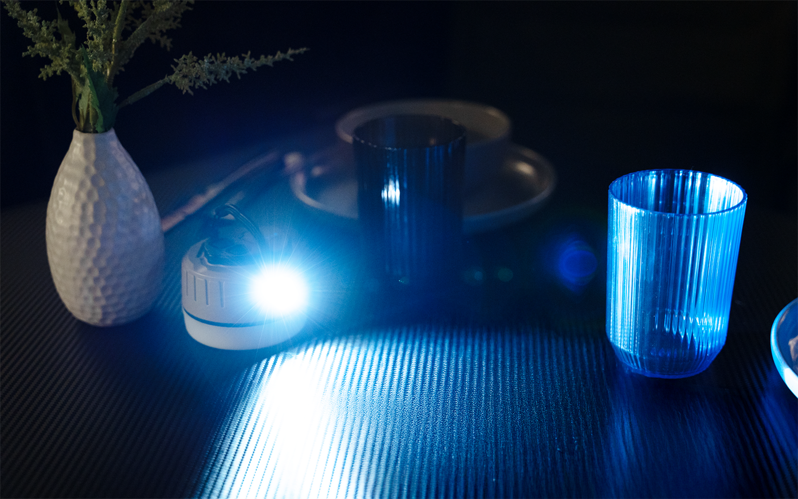 Kleines rundes Leuchtgerät strahlt starkes blaues Licht über eine geriffelte Oberfläche; rechts ein durchsichtiges blaues Trinkglas, links eine weiße Vase mit Pflanzstängeln, dahinter dunkle Teller