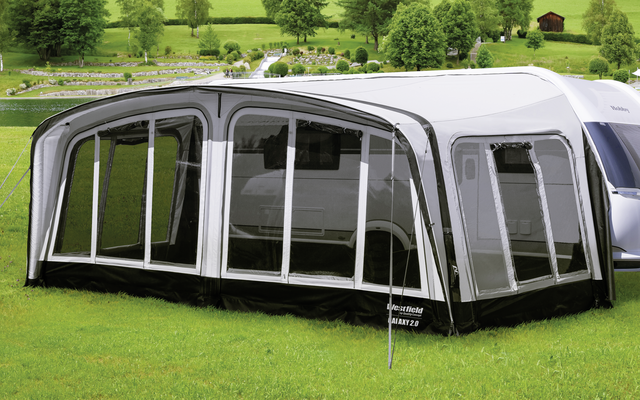 Caravane avec auvent fixé sur une prairie verte devant des collines boisées et des montagnes