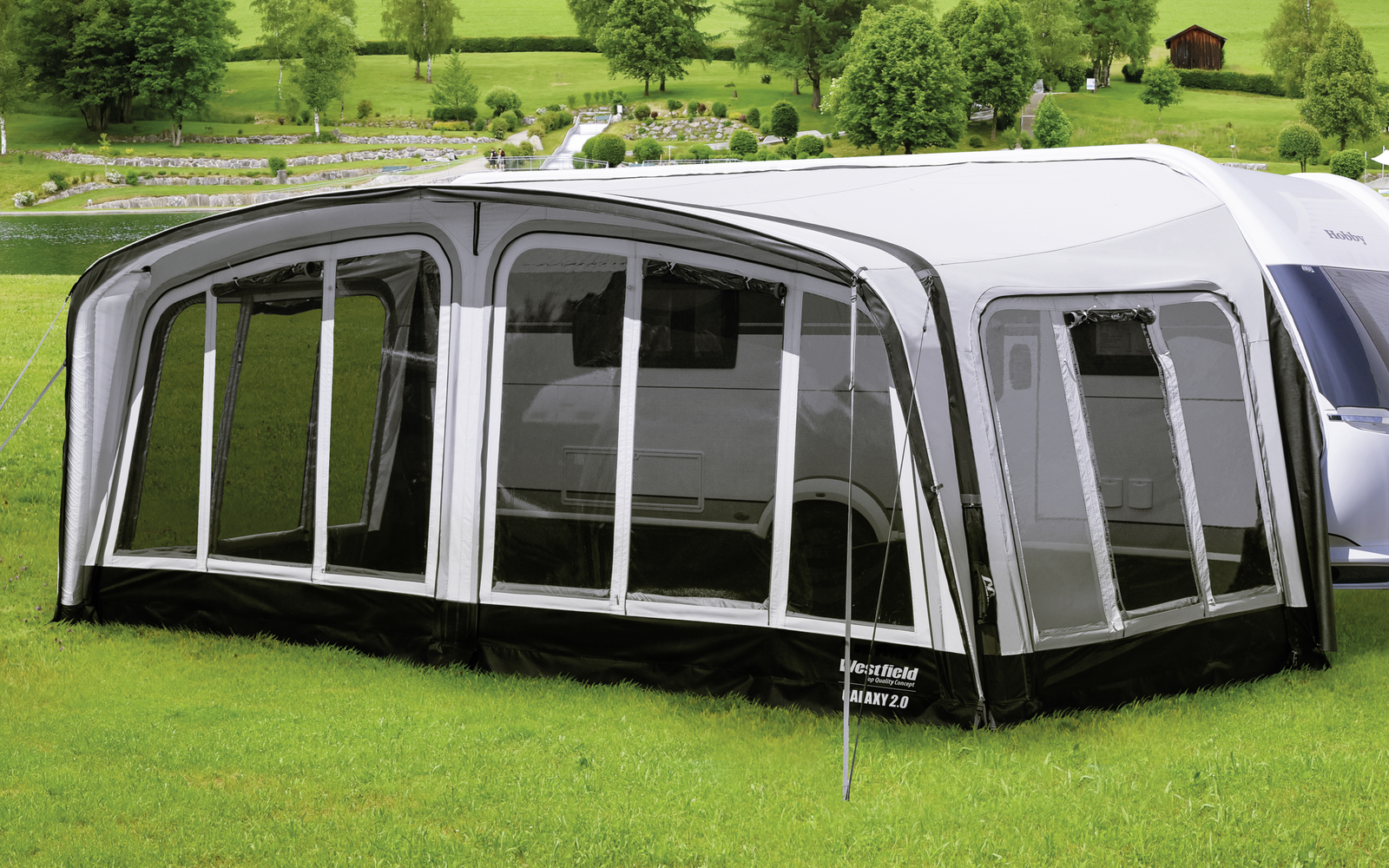 Caravane avec auvent fixé sur une prairie verte devant des collines boisées et des montagnes