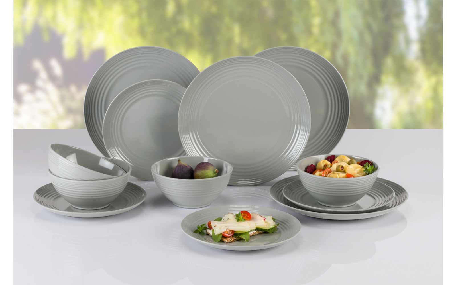 Mehrere hellgraue Teller und Schalen auf weißer Oberfläche; zwei Schalen mit dunklen Früchten, eine Schale mit gemischtem Obst, ein flacher Teller mit grünen Blättern, weißen Stücken und kleinen roten Tomaten. Verschwommener grüner Hintergrund