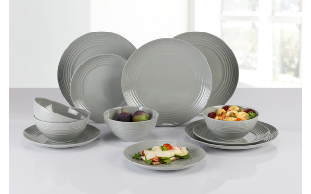 Graues Keramikgeschirr mit Tellern und Schalen auf weißer Oberfläche, einige Schalen mit Salat und Pflaumen, Fenster im Hintergrund