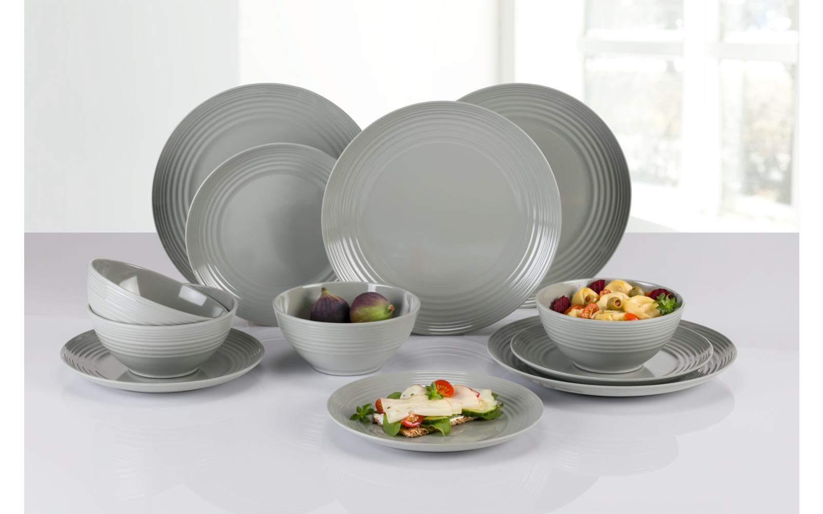 Graues Keramikgeschirr mit Tellern und Schalen auf weißer Oberfläche, einige Schalen mit Salat und Pflaumen, Fenster im Hintergrund