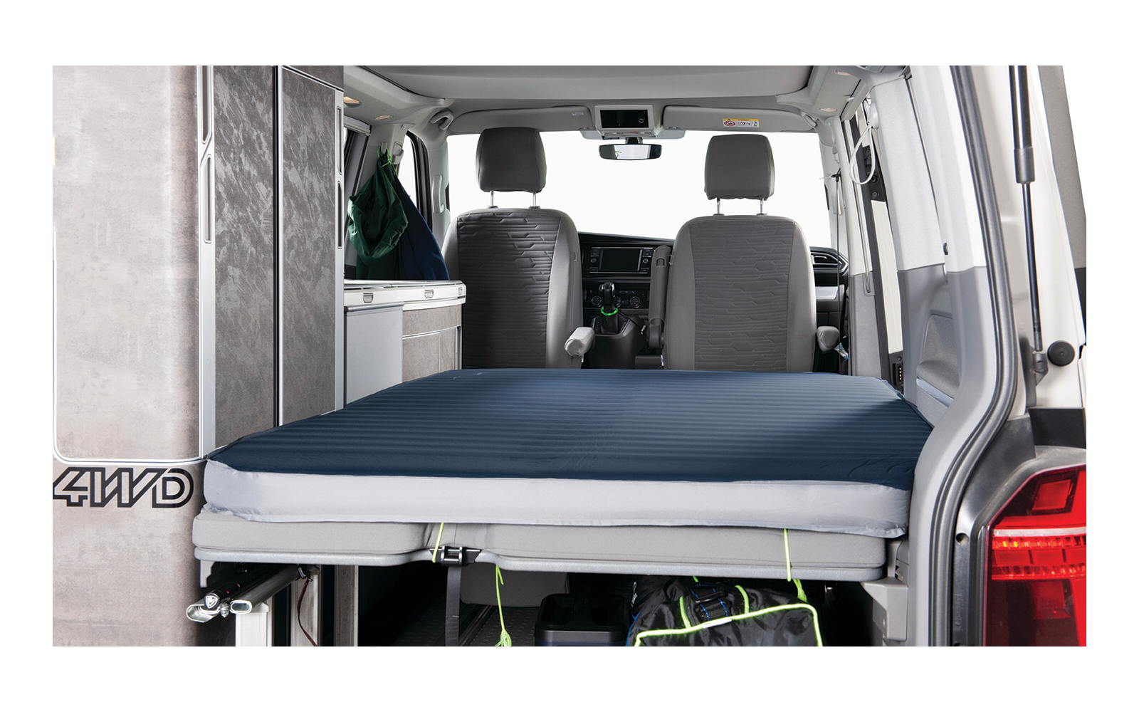 Interior of a van with extended bed: dark blue mattress on a platform, two front seats, cabinet on the left, bag under the platform, open rear door
