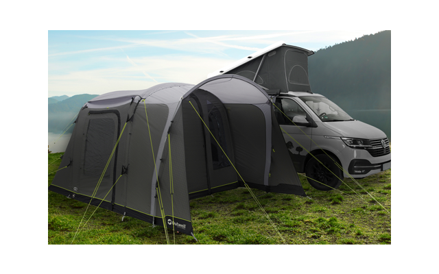 Wohnmobil mit angehängtem großen grauen Zelt auf einer Wiese neben See und Bergen