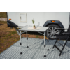 Table pliante avec petite plante en pot sur un napperon en dentelle et un gobelet blanc, deux chaises pliantes noires, roue et flanc d'une caravane en arrière-plan, tapis de sol à motif