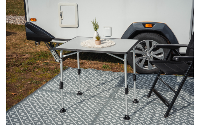 Table pliante avec petite plante en pot sur un napperon en dentelle et un gobelet blanc, deux chaises pliantes noires, roue et flanc d'une caravane en arrière-plan, tapis de sol à motif