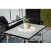 Table pliante avec napperon rond en crochet, vase blanc avec feuilles vertes effilées et petit pot beige; en arrière-plan la roue d’un véhicule blanc et une chaise noire sur un sol à motif