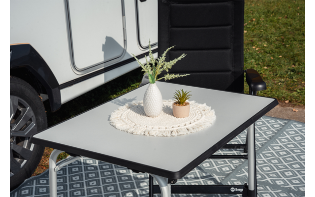 Table pliante avec napperon rond en crochet, vase blanc avec feuilles vertes effilées et petit pot beige; en arrière-plan la roue d’un véhicule blanc et une chaise noire sur un sol à motif