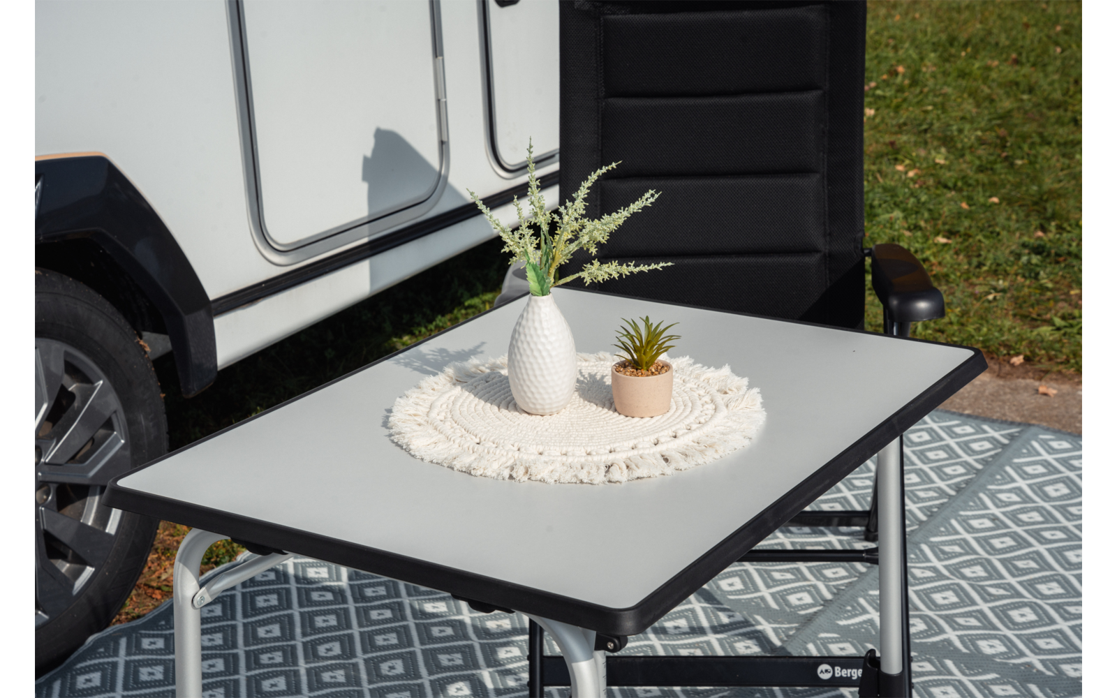 Table pliante avec napperon rond en crochet, vase blanc avec feuilles vertes effilées et petit pot beige; en arrière-plan la roue d’un véhicule blanc et une chaise noire sur un sol à motif