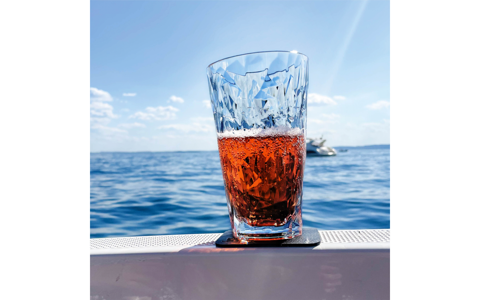 Geschliffenes Glas mit rötlich-orangenem Getränk auf Untersetzer auf der Reling eines Bootes, dahinter Meer, Himmel und kleines Motorboot in der Ferne