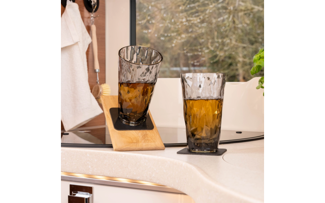 Zwei getönte, facettierte Trinkgläser mit brauner Flüssigkeit auf schwarzen Untersetzern auf einer hellen Arbeitsplatte; ein Glas steht schräg auf einem Holzbrett. Im Hintergrund ein Fenster mit Bäumen, links ein Handtuch und eine Bürste, rechts eine