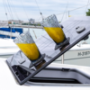 Zwei geschliffene Gläser mit orangener Flüssigkeit auf einer schrägen, glänzenden schwarzen Platte auf einem weißen Boot, im Hintergrund Segelboote und Wasser