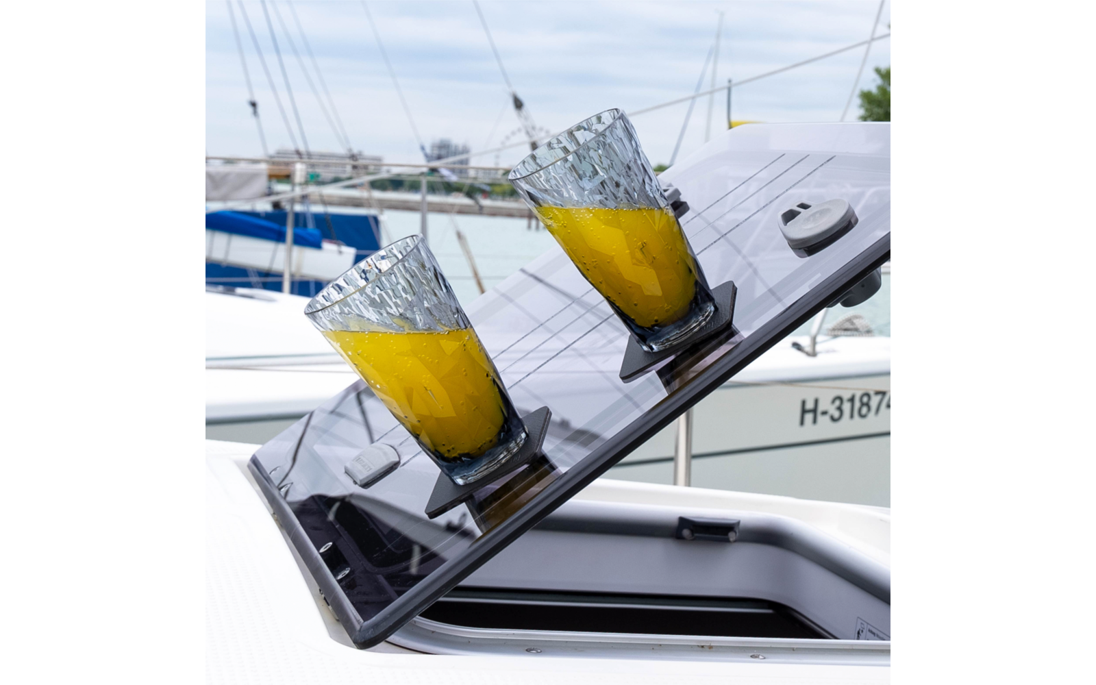 Zwei geschliffene Gläser mit orangener Flüssigkeit auf einer schrägen, glänzenden schwarzen Platte auf einem weißen Boot, im Hintergrund Segelboote und Wasser