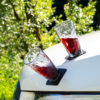 Zwei facettierte Gläser mit roter Flüssigkeit, schräg auf schwarzen Untersetzern auf der weißen Motorhaube eines Autos, unscharfer grüner Hintergrund