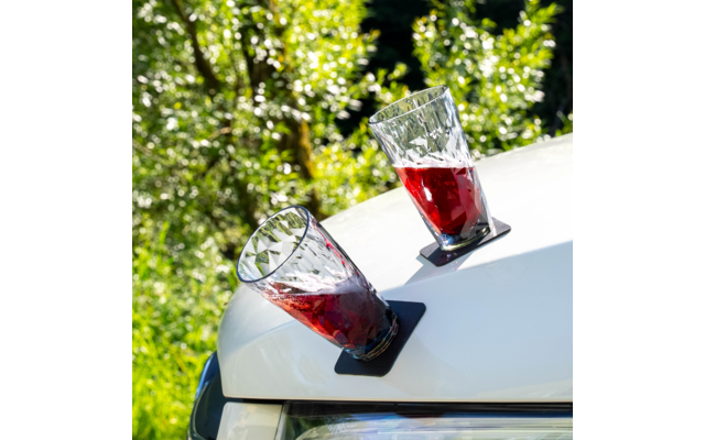 Zwei facettierte Gläser mit roter Flüssigkeit, schräg auf schwarzen Untersetzern auf der weißen Motorhaube eines Autos, unscharfer grüner Hintergrund