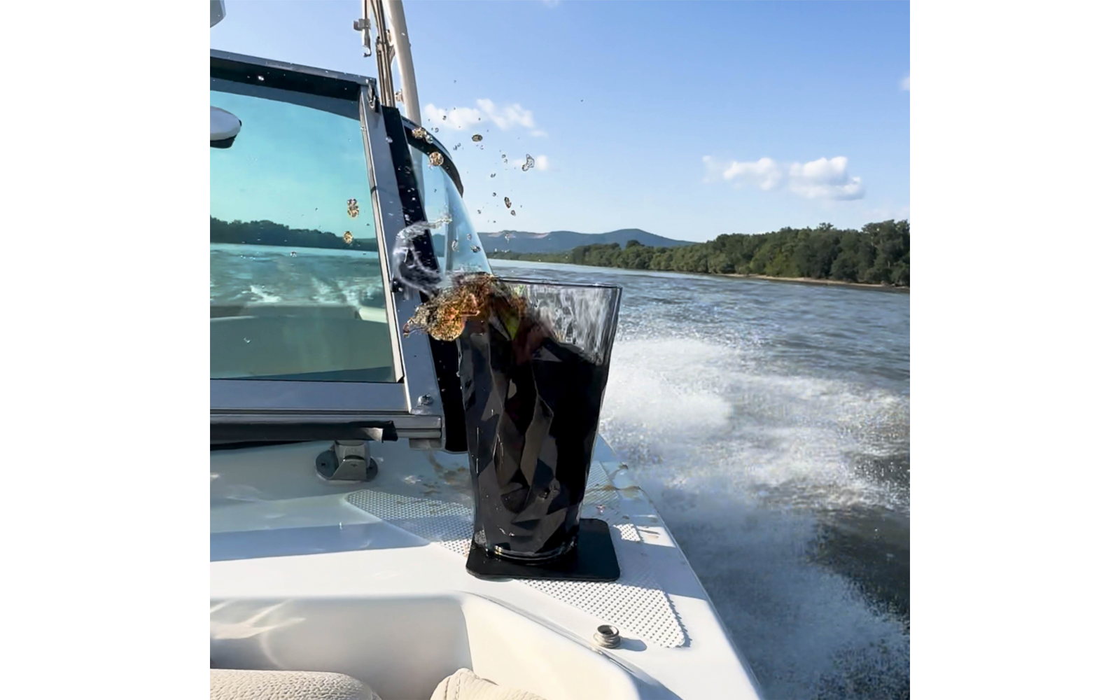 Dunkles Getränk spritzt aus einem hohen Glas auf dem weißen Bootsdeck; Bootsfenster, Heckwelle auf dem See, bewaldete Ufer, Hügel und blauer Himmel mit Wolken