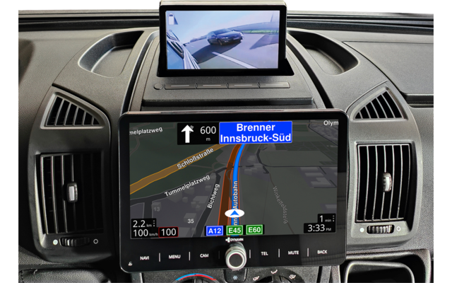 Armaturenbrett eines Fahrzeugs mit zwei Bildschirmen: großer Bildschirm zeigt Karte mit blau markierter Route und Schriftzug "Brenner Innsbruck-Süd", kleiner oberer Bildschirm zeigt Rückfahrkamera mit Straße und einem Auto; seitliche Lüftungsdüsen un