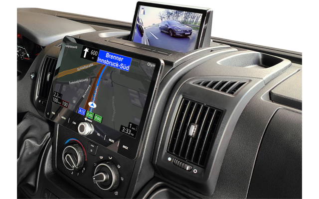 Fahrzeug-Mittelkonsole mit zwei Bildschirmen: großer Touchscreen zeigt Navigationskarte mit Route und dem Text 'Brenner Innsbruck-Süd', kleiner ausklappbarer Monitor zeigt Kamerabild einer Straße mit einem Auto; darunter Klimadrehregler, rechteckiger