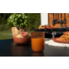 Glas mit orangefarbenem Saft auf schwarzem Tisch, daneben Schale mit Pfirsichen, Basilikum in Topf, Teller mit Croissants und ein Grill im Hintergrund