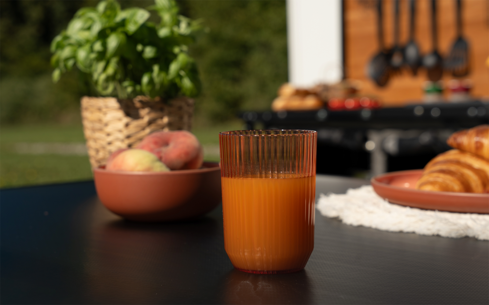 Glas mit orangefarbenem Saft auf schwarzem Tisch, daneben Schale mit Pfirsichen, Basilikum in Topf, Teller mit Croissants und ein Grill im Hintergrund