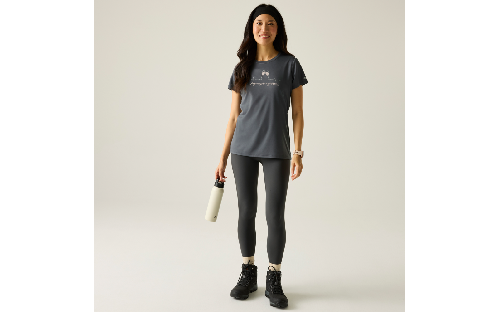 Femme en t‑shirt de sport foncé, legging noir et bottines, portant un bonnet noir, tenant une gourde claire dans la main droite, debout sur fond clair