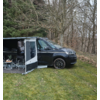 Schwarzer Campingbus mit ausgefahrenem Vorzelt, geöffneter Seitentür, Campingstühlen und Tisch auf Gras vor kahlen Bäumen