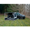 Schwarzer Camper mit ausgefahrener Markise und seitlichem Wandteil, zwei Campingstühle und kleiner Tisch auf einer Bodenmatte, geparkt auf Gras vor kahlen Bäumen