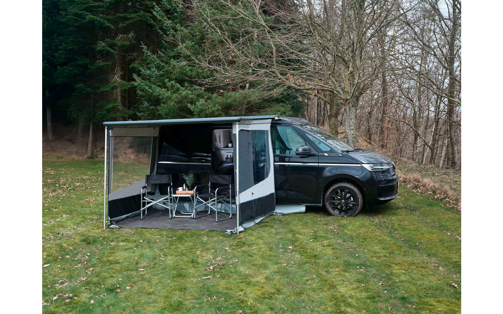 Schwarzer Camper mit ausgefahrener Markise und seitlichem Wandteil, zwei Campingstühle und kleiner Tisch auf einer Bodenmatte, geparkt auf Gras vor kahlen Bäumen