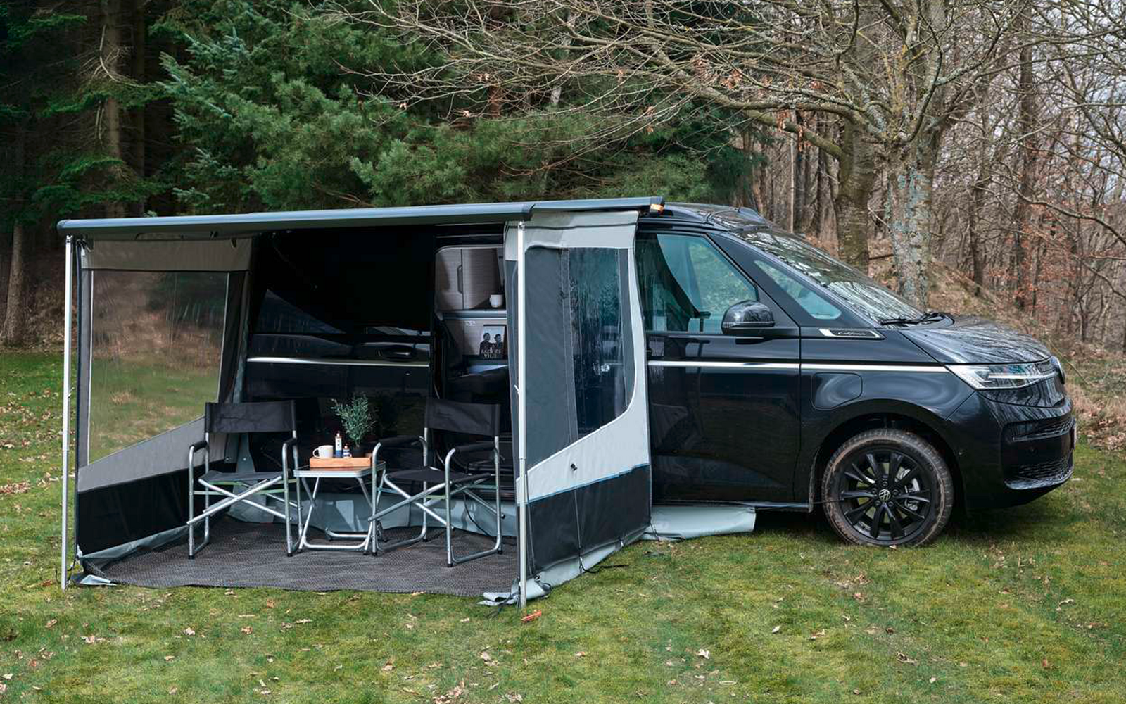 Schwarzer Camper mit ausgefahrener Markise und seitlichem Wandteil, zwei Campingstühle und kleiner Tisch auf einer Bodenmatte, geparkt auf Gras vor kahlen Bäumen