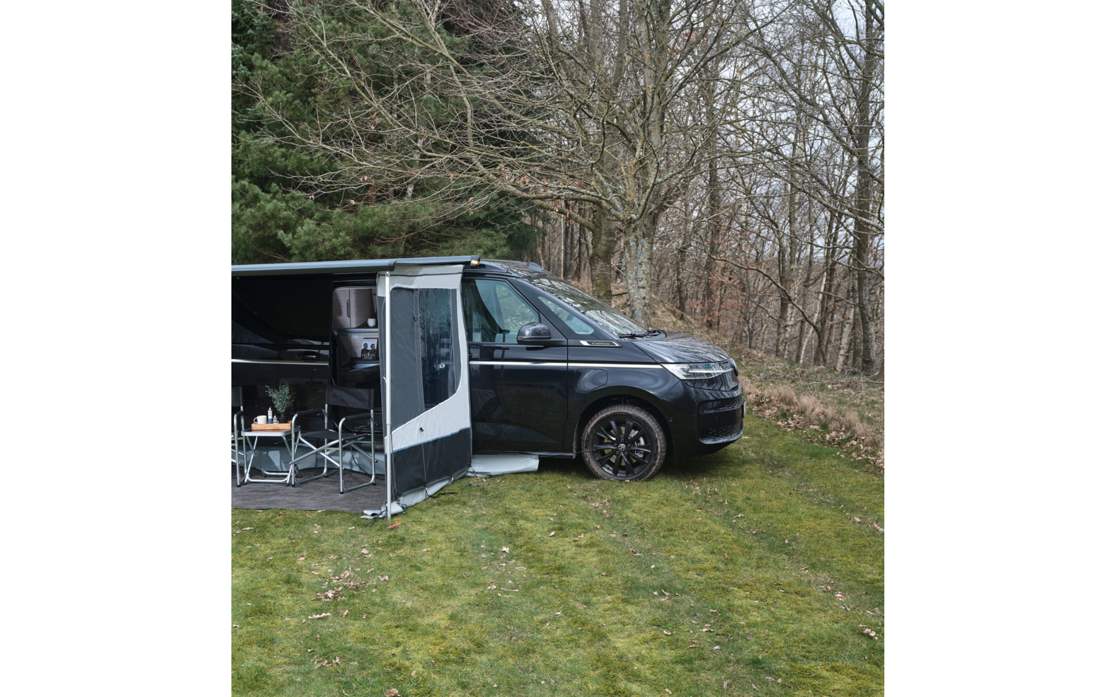 Schwarzer Campingbus mit ausgefahrenem Vorzelt, geöffneter Seitentür, Campingstühlen und Tisch auf Gras vor kahlen Bäumen