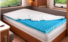 Ein Einzelbett in einem Wohnmobil mit weißer gesteppter Auflage teilweise zurückgefaltet, blauem Noppen-Schaumstofftopper, Holzverkleidung, zwei Fenstern mit Gardinen und Blick auf grüne Pflanzen
