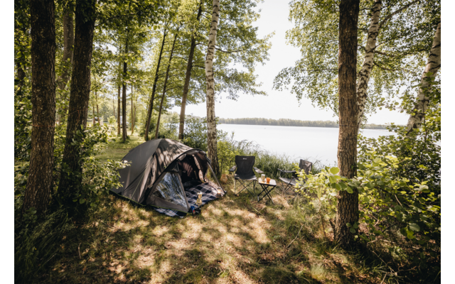 Zelt mit offenem Eingang, Campingstühle und kleiner Tisch unter Bäumen am Ufer eines Sees