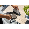 Deux mains : l'une tient une petite assiette ronde brune, l'autre une brosse à main beige et balaie des miettes sur une table en bois clair ; à droite un pot de plante blanc avec des feuilles vertes ; à gauche une partie d'une chaise avec un coussin bleu