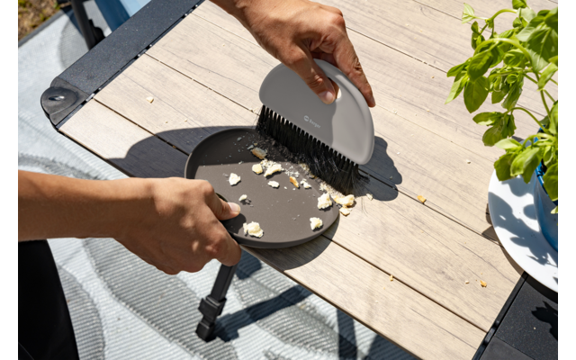 Deux mains : l'une tient une petite assiette ronde brune, l'autre une brosse à main beige et balaie des miettes sur une table en bois clair ; à droite un pot de plante blanc avec des feuilles vertes ; à gauche une partie d'une chaise avec un coussin bleu