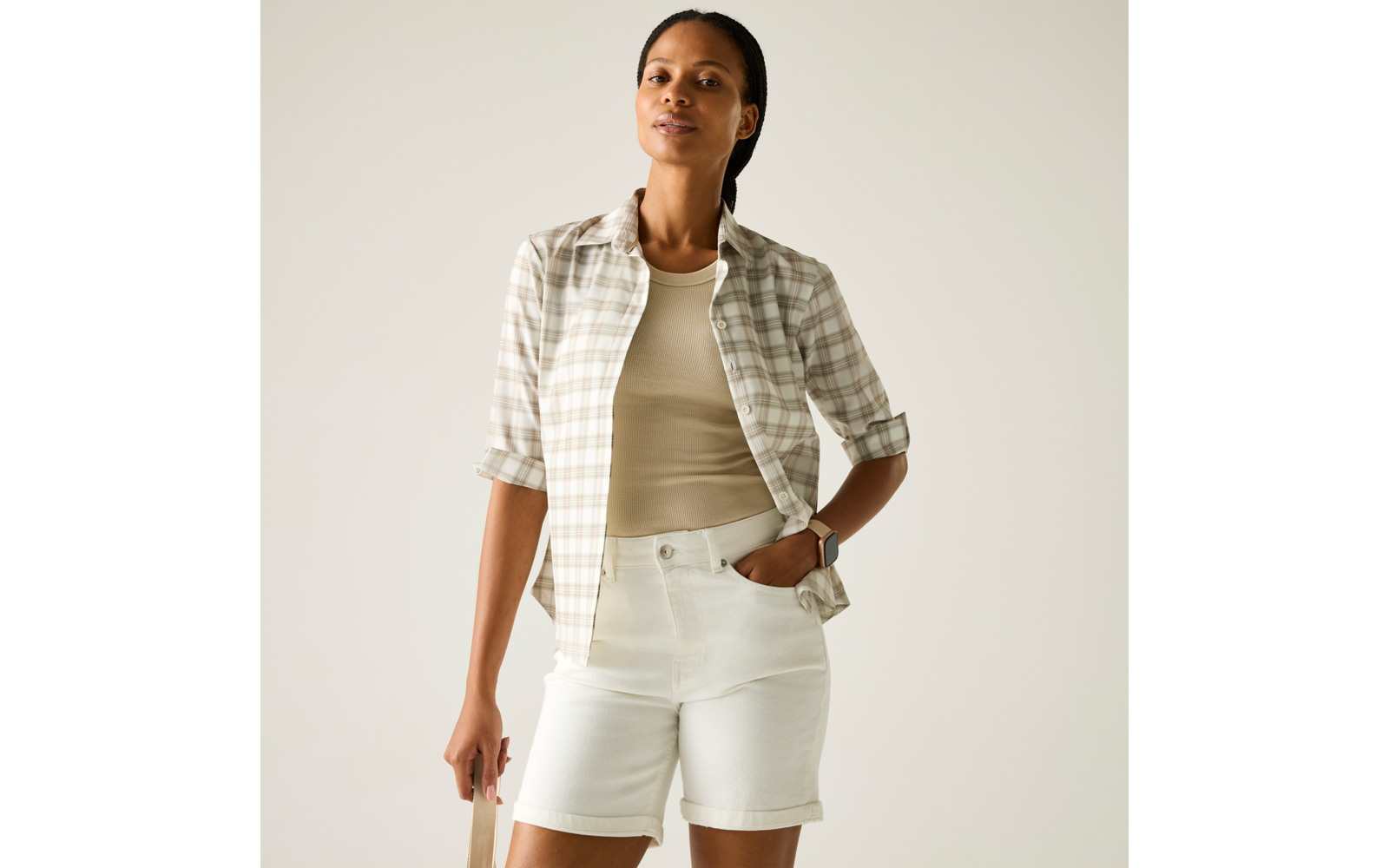 Person vor hellem Hintergrund, trägt beige Tanktop, hell karierte Hemdjacke mit hochgekrempelten Ärmeln, weiße Shorts; eine Hand in der Hosentasche, hält eine beige Tasche