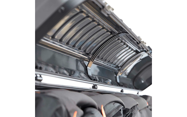 Nahaufnahme eines geöffneten metallischen Gehäuses mit gebogenen Lamellen und glänzenden Metallstäben, darunter schwarze Jacken mit orangefarbenen Details