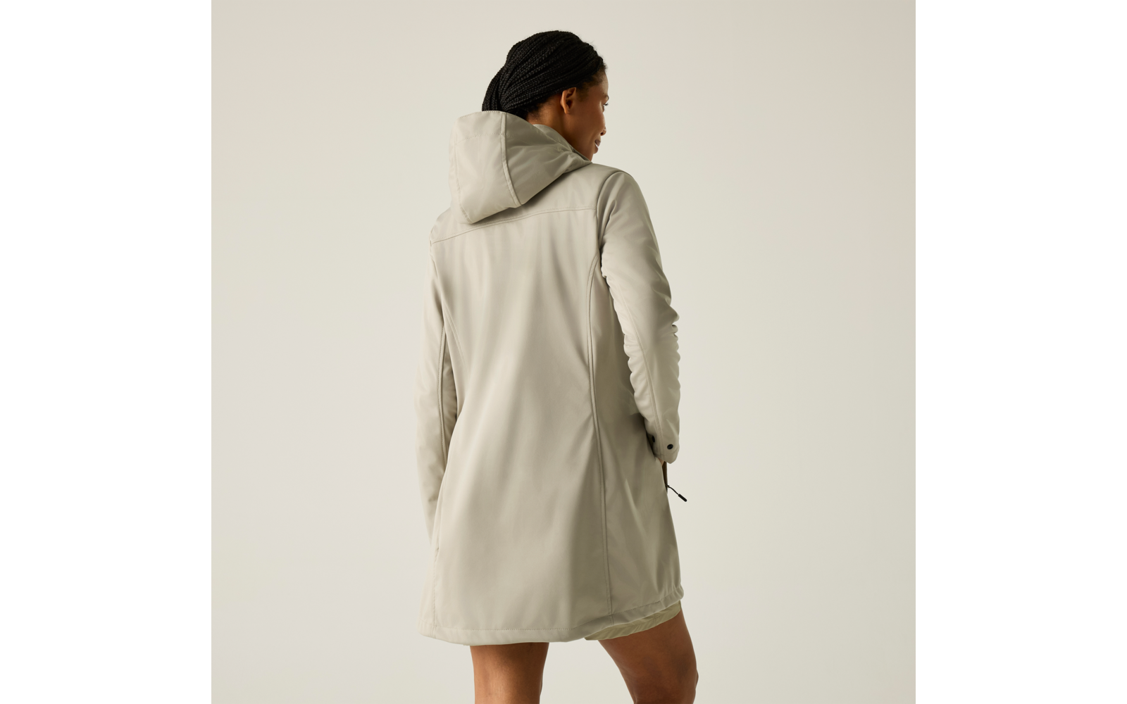 Rückansicht einer Person mit geflochtenen Haaren, die einen hellbeigen Kapuzenmantel trägt, vor einfarbig hellem Hintergrund