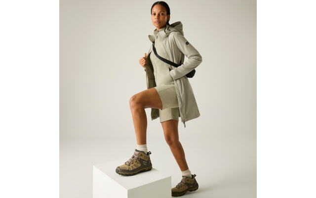 Person in heller Kapuzenjacke und Shorts, eine Hand in der Jacke, ein Fuß auf einem weißen Würfel, trägt Trekking-Schuhe und eine kleine Umhängetasche vor neutralem Hintergrund