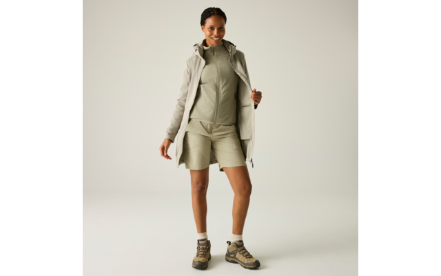 Person in hellgrüner Zip-Jacke und passenden Shorts, offener beiger Mantel, Knöchelsocken und Wanderschuhe, steht vor neutralem Hintergrund
