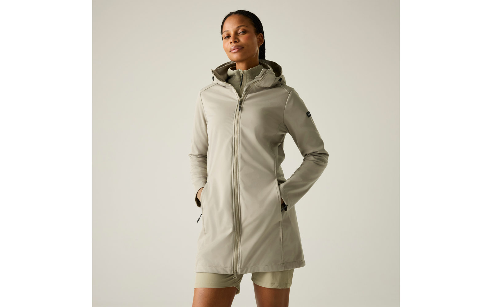 Frau steht vor hellem, einfarbigem Hintergrund, trägt einen hellolivgrünen langen Kapuzenmantel mit durchgehendem Reißverschluss und seitlichen Taschen, Hände in den Taschen, kurze Hose darunter sichtbar