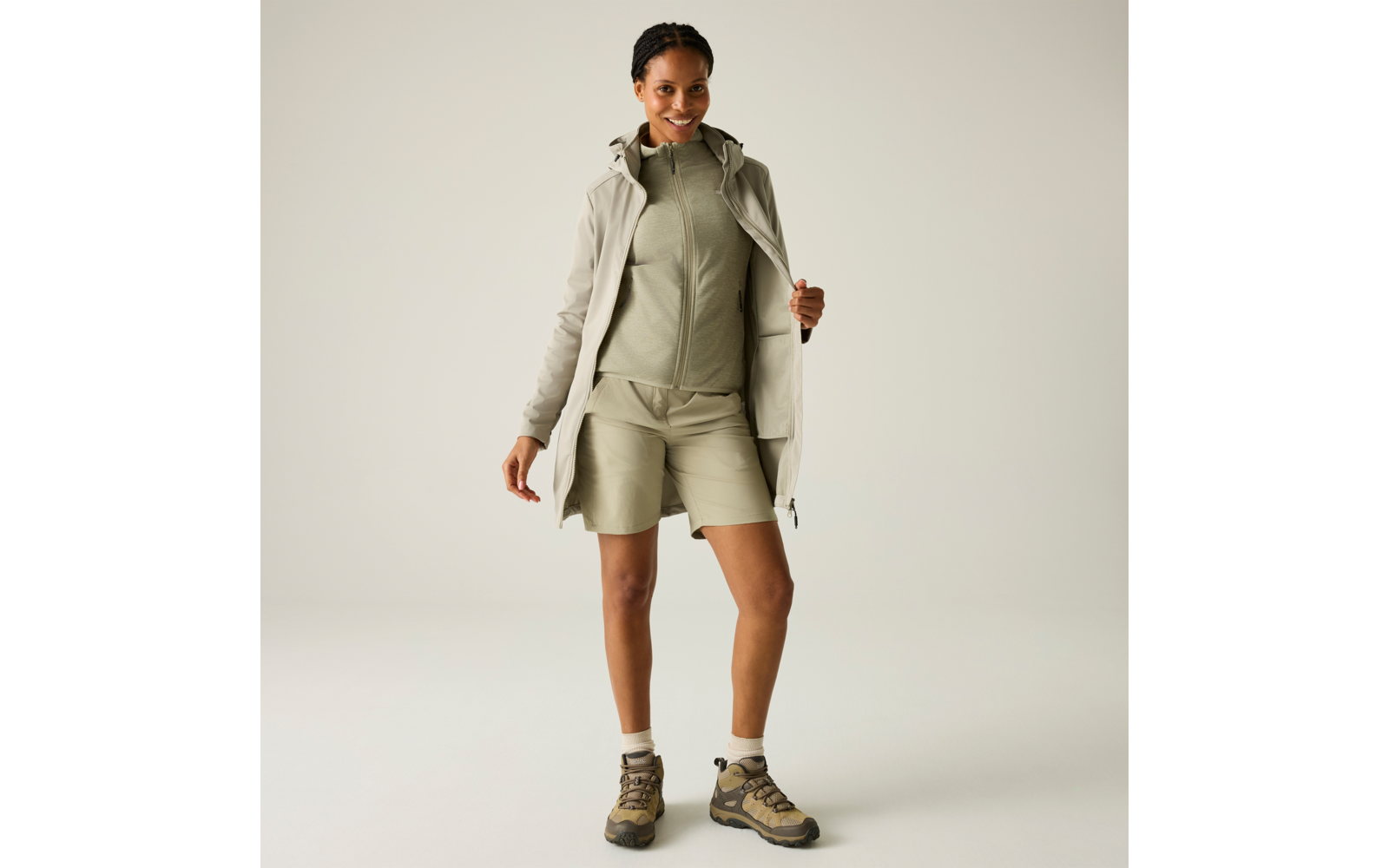 Person in hellgrüner Zip-Jacke und passenden Shorts, offener beiger Mantel, Knöchelsocken und Wanderschuhe, steht vor neutralem Hintergrund