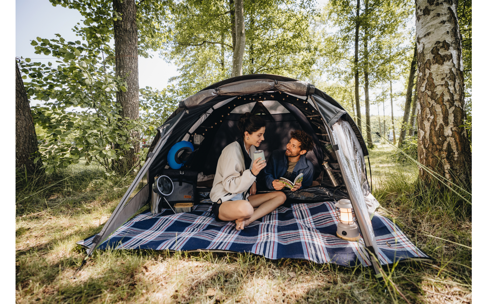 Zwei Personen sitzen in einem Zelt auf einer karierten Decke; eine hält eine Tasse, die andere ein aufgeschlagenes Buch; Zelt steht zwischen Bäumen, Laterne und Rucksack daneben