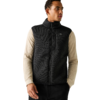 Persoon draagt beige shirt met lange mouwen onder een zwarte mouwloze ritsvest, staat voor een lichte achtergrond, handen in de zakken, kijkt naar rechts