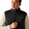Bovenlichaam van een persoon met een zwarte fleece-bodywarmer met rits en borstzak over een beige longsleeve, houdt de rits vast, kijkt naar rechts