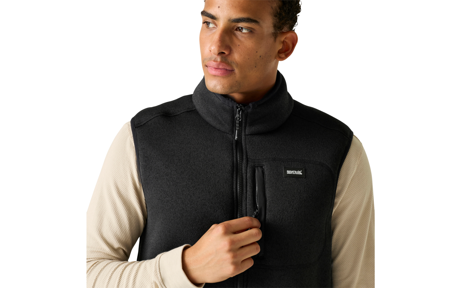 Bovenlichaam van een persoon met een zwarte fleece-bodywarmer met rits en borstzak over een beige longsleeve, houdt de rits vast, kijkt naar rechts