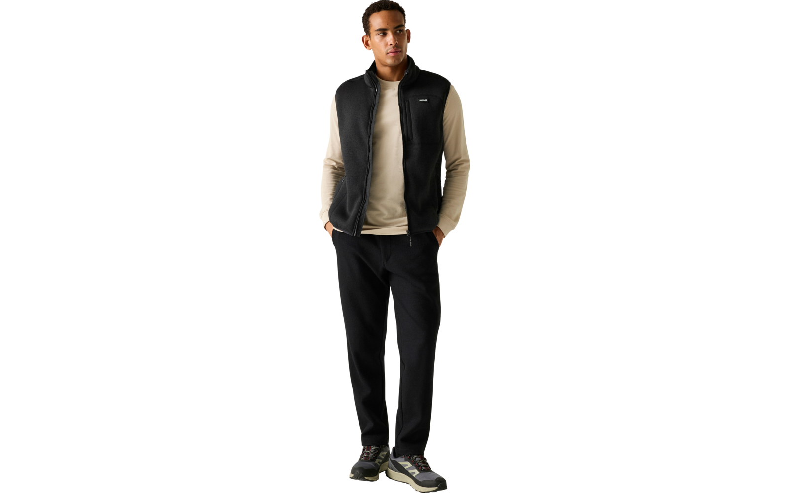 Een staande man met handen in zijn zakken, draagt een beige longsleeve, een zwart mouwloos ritsvest, zwarte broek en sneakers voor een effen lichtgrijze achtergrond