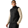 Persoon voor een lichtgrijze achtergrond, draagt een zwarte mouwloze ritssluiting-vest over een beige longsleeve, één hand in de zak, kijkt naar rechts