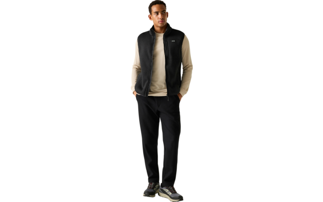 Een staande man met handen in zijn zakken, draagt een beige longsleeve, een zwart mouwloos ritsvest, zwarte broek en sneakers voor een effen lichtgrijze achtergrond