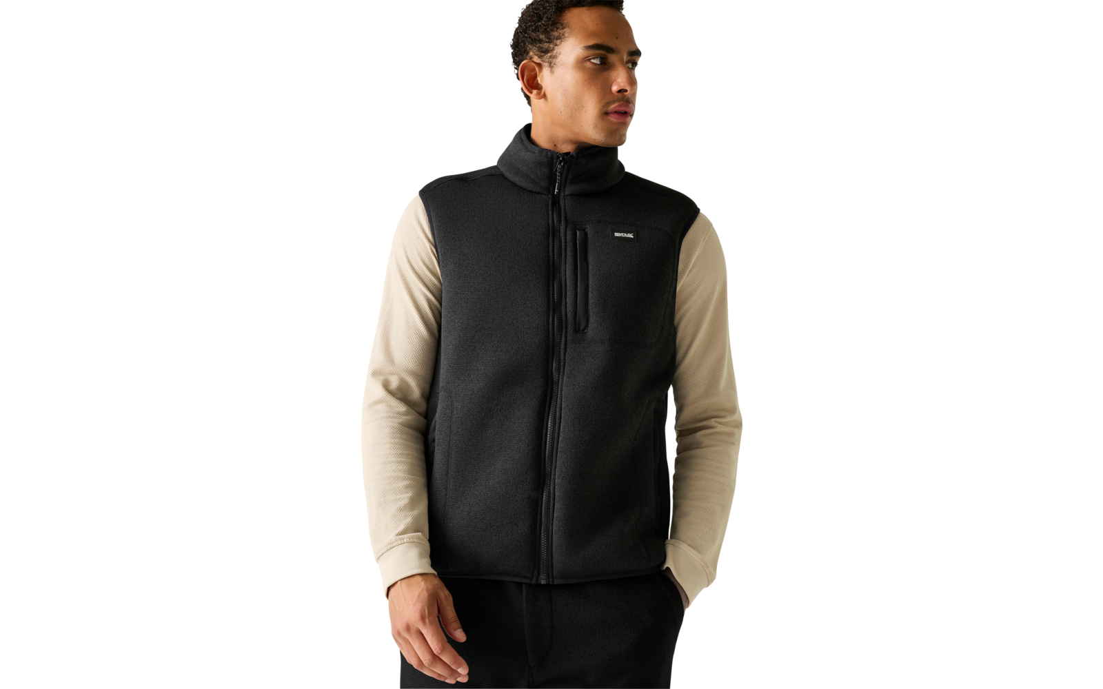 Persoon draagt beige shirt met lange mouwen onder een zwarte mouwloze ritsvest, staat voor een lichte achtergrond, handen in de zakken, kijkt naar rechts